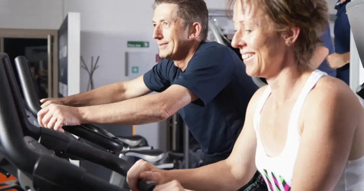 Uśmiechnięci ludzie ćwiczą na rowerach stacjonarnych. Czy ćwiczyć codziennie? Oni chyba tak robią, bo wyglądają na zadowolonych.