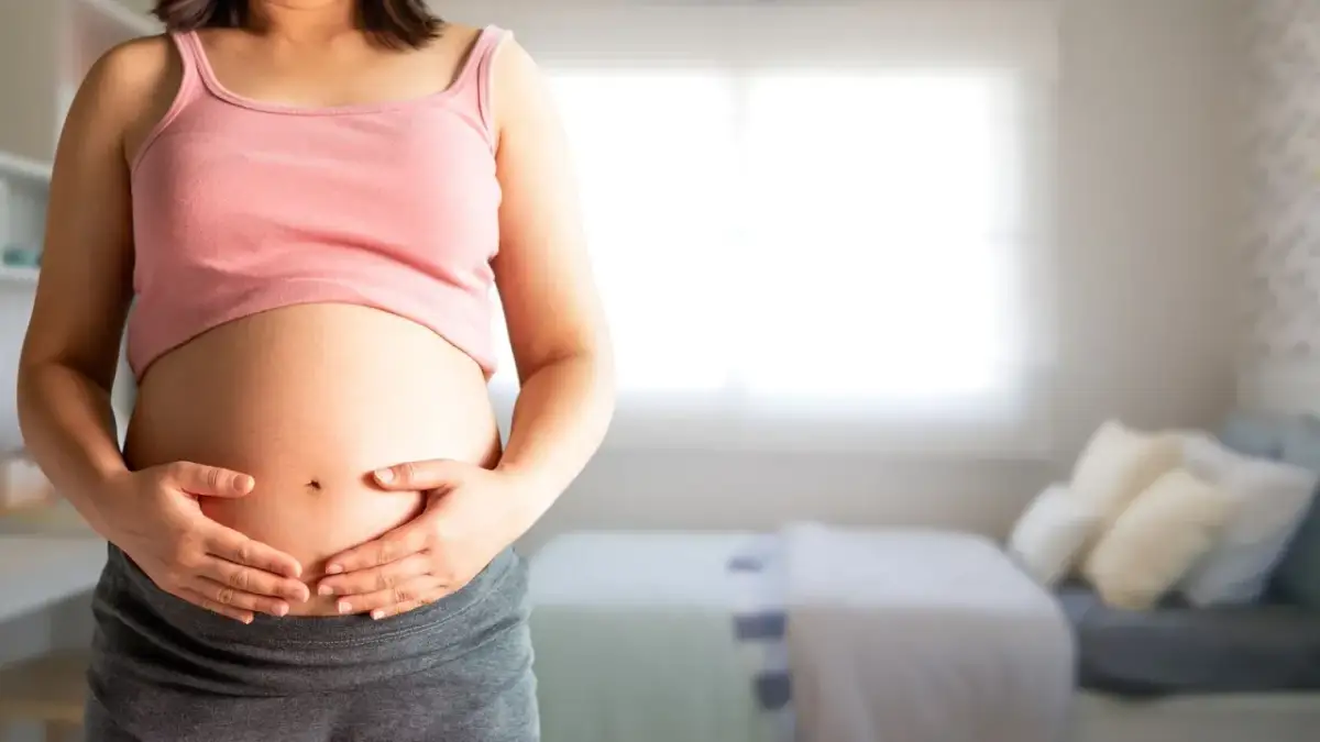Brzuch kobiety w ciąży, trzymany dłońmi, na tle pokoju.
