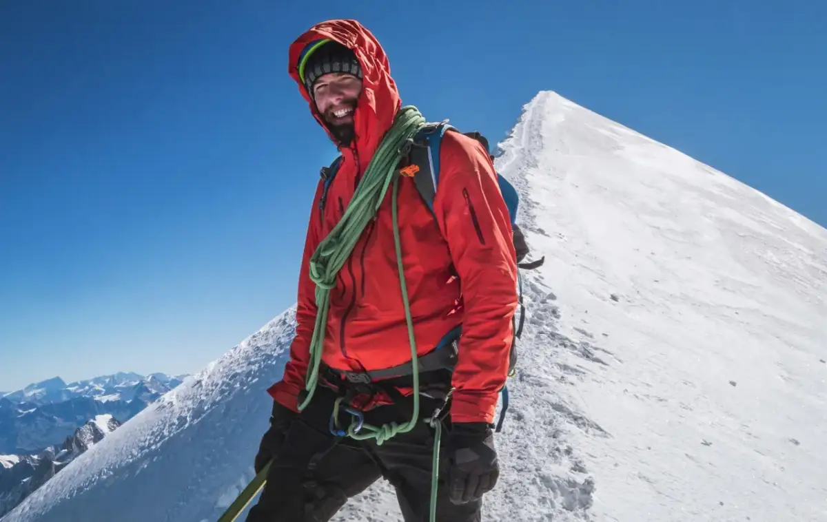 Uśmiechnięty alpinista na szczycie Mont Blanc, w pomarańczowej kurtce i z liną, cieszy się widokiem Alp.