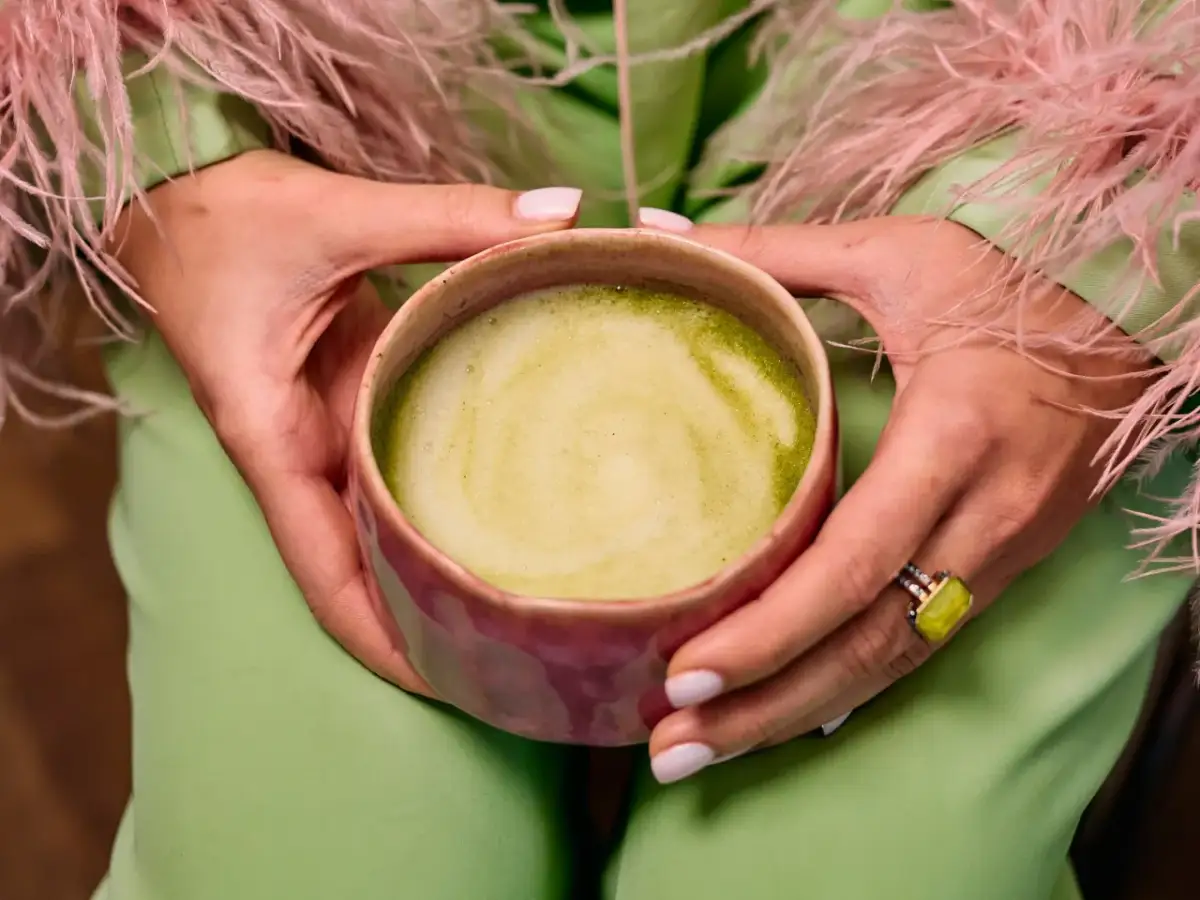 Dłonie trzymają kubek z zieloną matchą, zastanawiając się, czy matcha odchudza. Ubrana w pistacjowy strój z różowymi piórami.