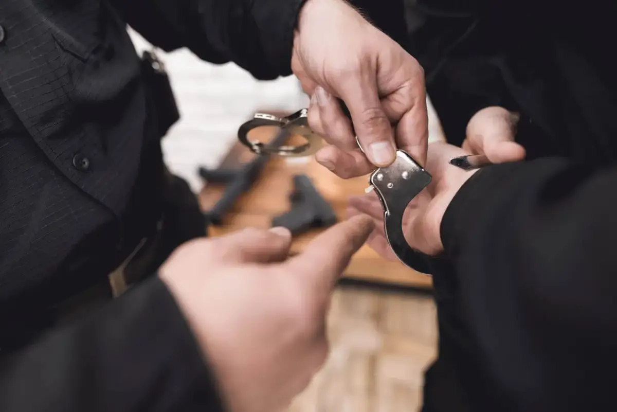 Dłoń policjanta zakłada kajdanki. Czy policjant po służbie może wystawić mandat? Pytanie pozostaje otwarte, gdy widzimy broń na stole.