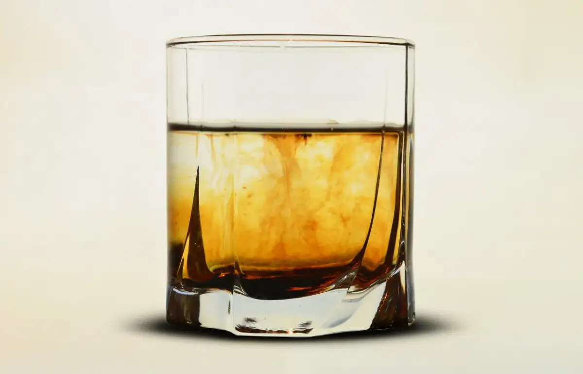 Szklanka z bursztynowym płynem, w którym rozpuszcza się ciemniejsza substancja, przypominająca proces, jak zrobic whiskey.