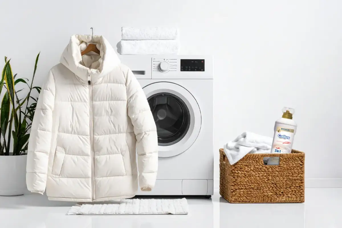 Biała kurtka zimowa wisi obok pralki, gotowa do prania. Obok koszyk z ręcznikami i płynem do prania. Dowiedz się, jak prać kurtkę zimową w pralce.