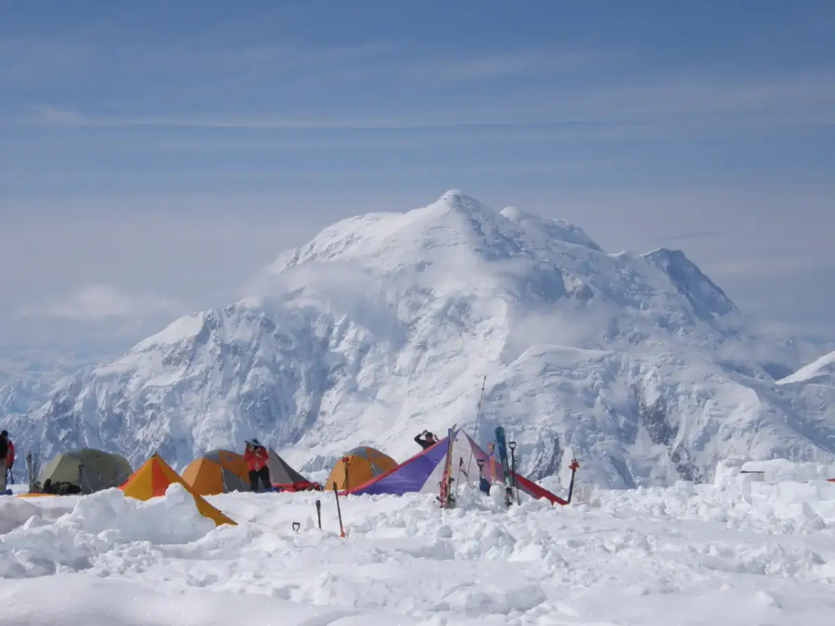 Namiotowe miasteczko u stóp McKinley szczyt. W tle majestatyczna, ośnieżona góra, a na pierwszym planie kolorowe namioty i sprzęt narciarski.