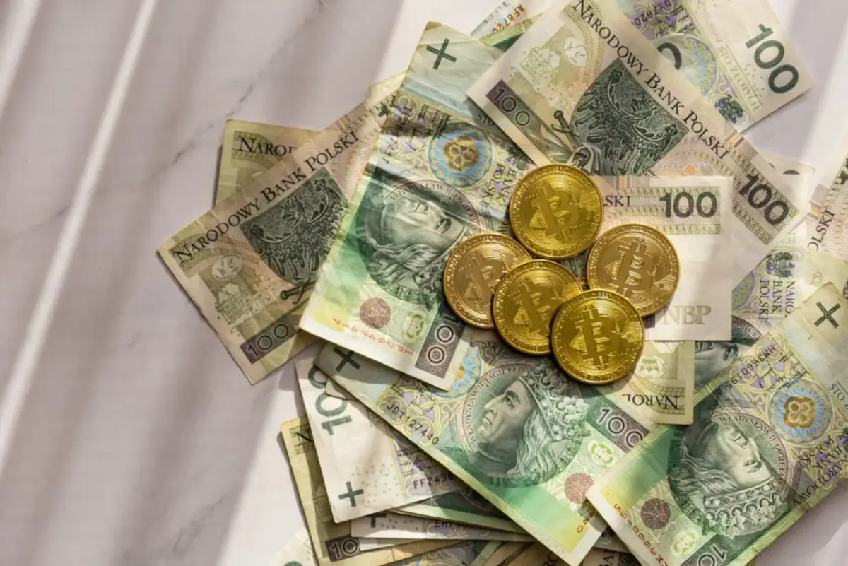 Złote monety z symbolem Bitcoina leżą na stosie polskich banknotów 100 zł. Czy w kantorze można rozmienić pieniądze?