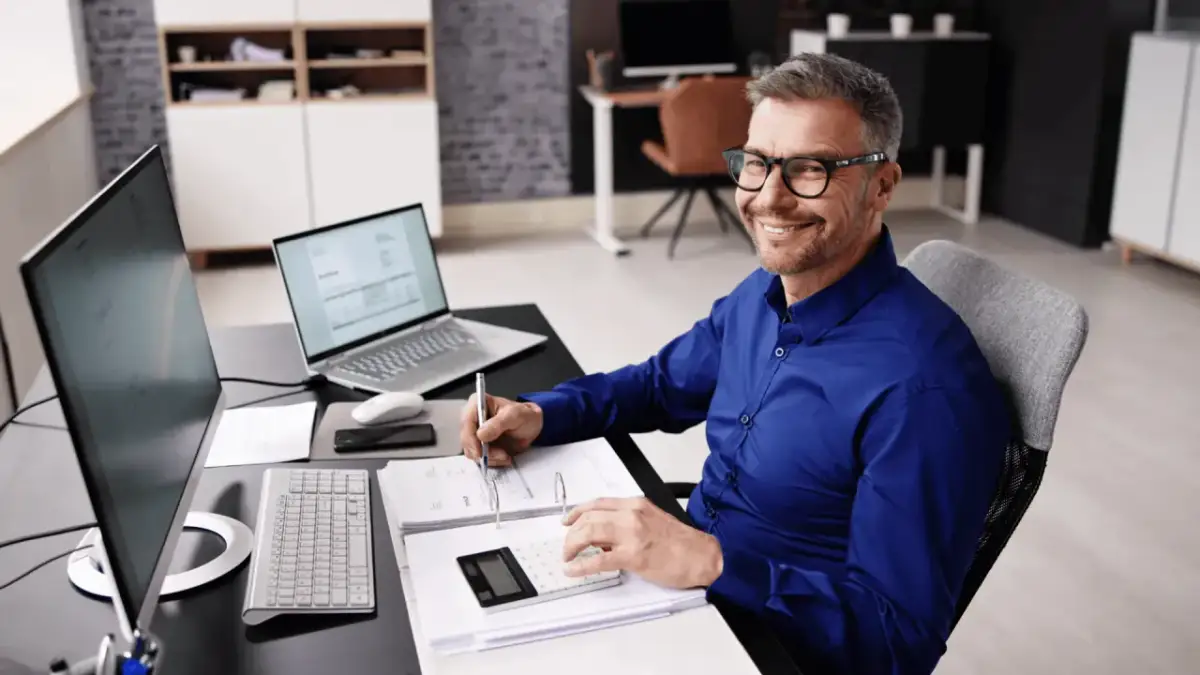 Uśmiechnięty mężczyzna w okularach przy biurku z komputerem i dokumentami. Zastanawia się, ile można dostać dofinansowania na firmę.