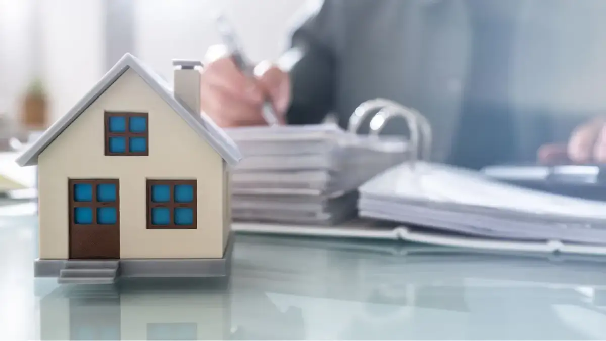 Model domu na biurku, obok stosy dokumentów i osoba pisząca. Czy kupując dom od dewelopera płacę podatek?