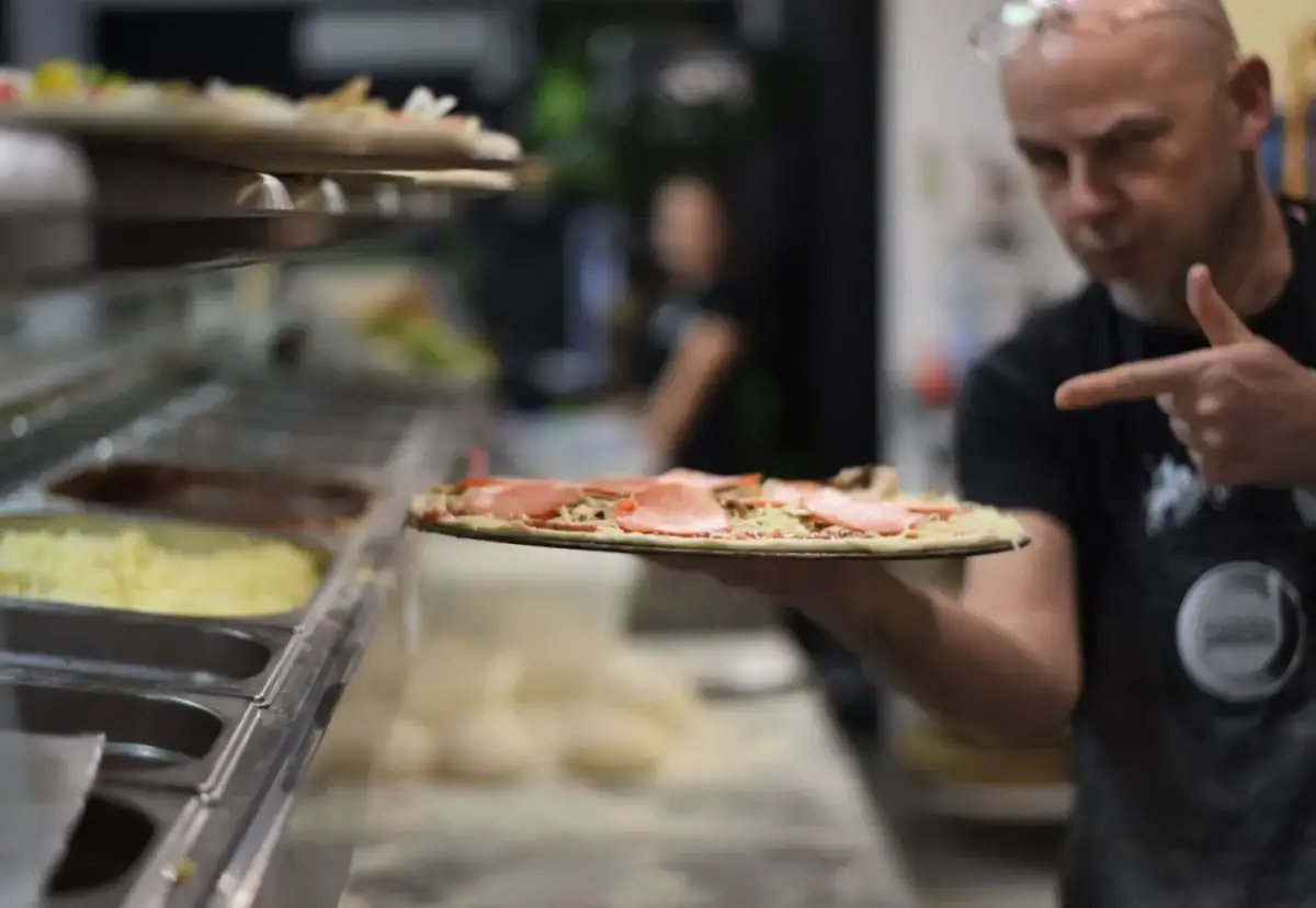 Mężczyzna w fartuchu pokazuje surową pizzę z szynką i pieczarkami.