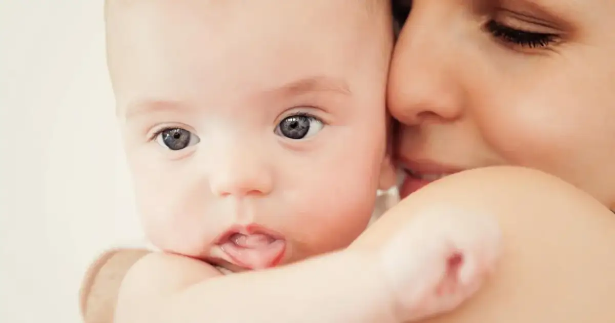 Zbliżenie na twarz niemowlaka z wyciągniętym językiem, przytulonego do matki.