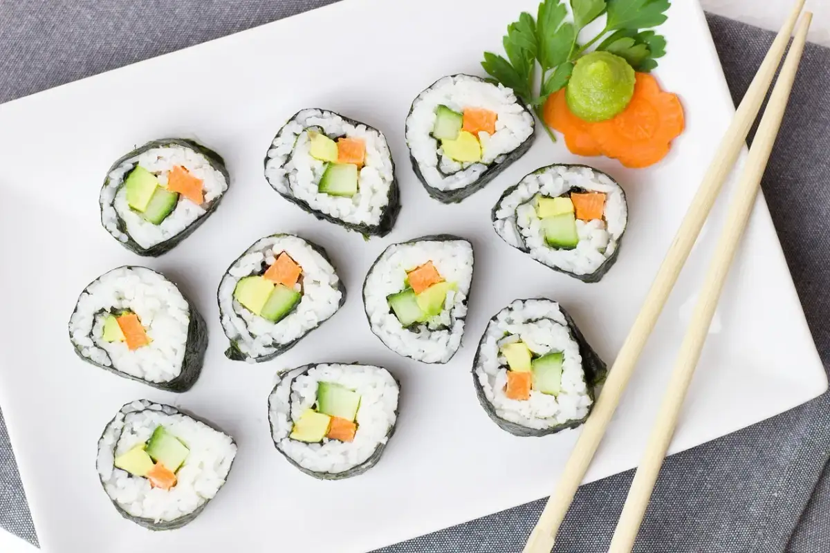 Talerz z rolkami sushi z awokado, ogórkiem i marchewką. Obok zielona pasta wasabi i ozdobna marchewka. Pałeczki czekają, by zacząć ucztę z czym sushi.