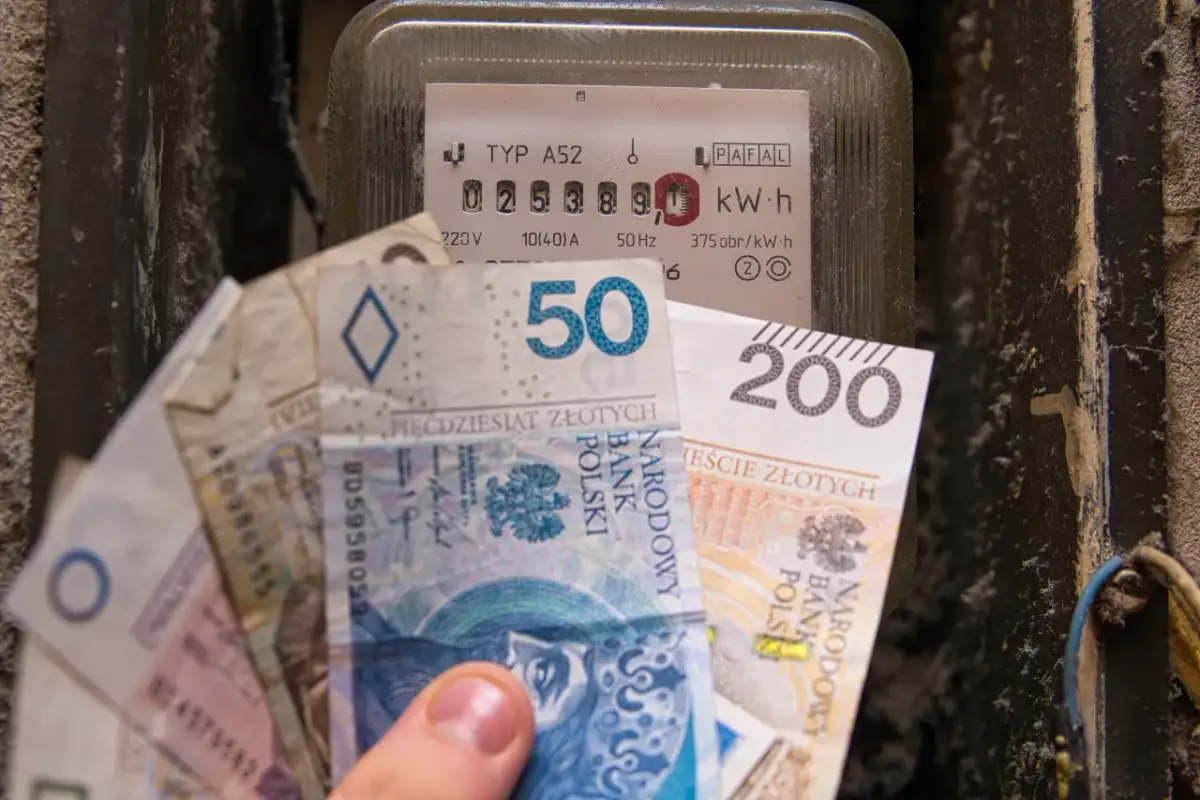 Dłoń trzyma banknoty obok licznika energii, symbolizując rosnące ceny energii elektrycznej.