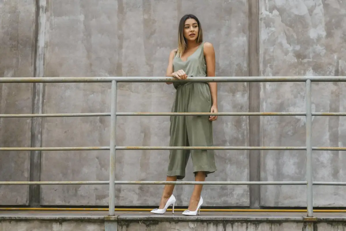 Kobieta w zielonym kombinezonie i białych szpilkach stoi przy metalowej balustradzie na tle betonowej ściany.