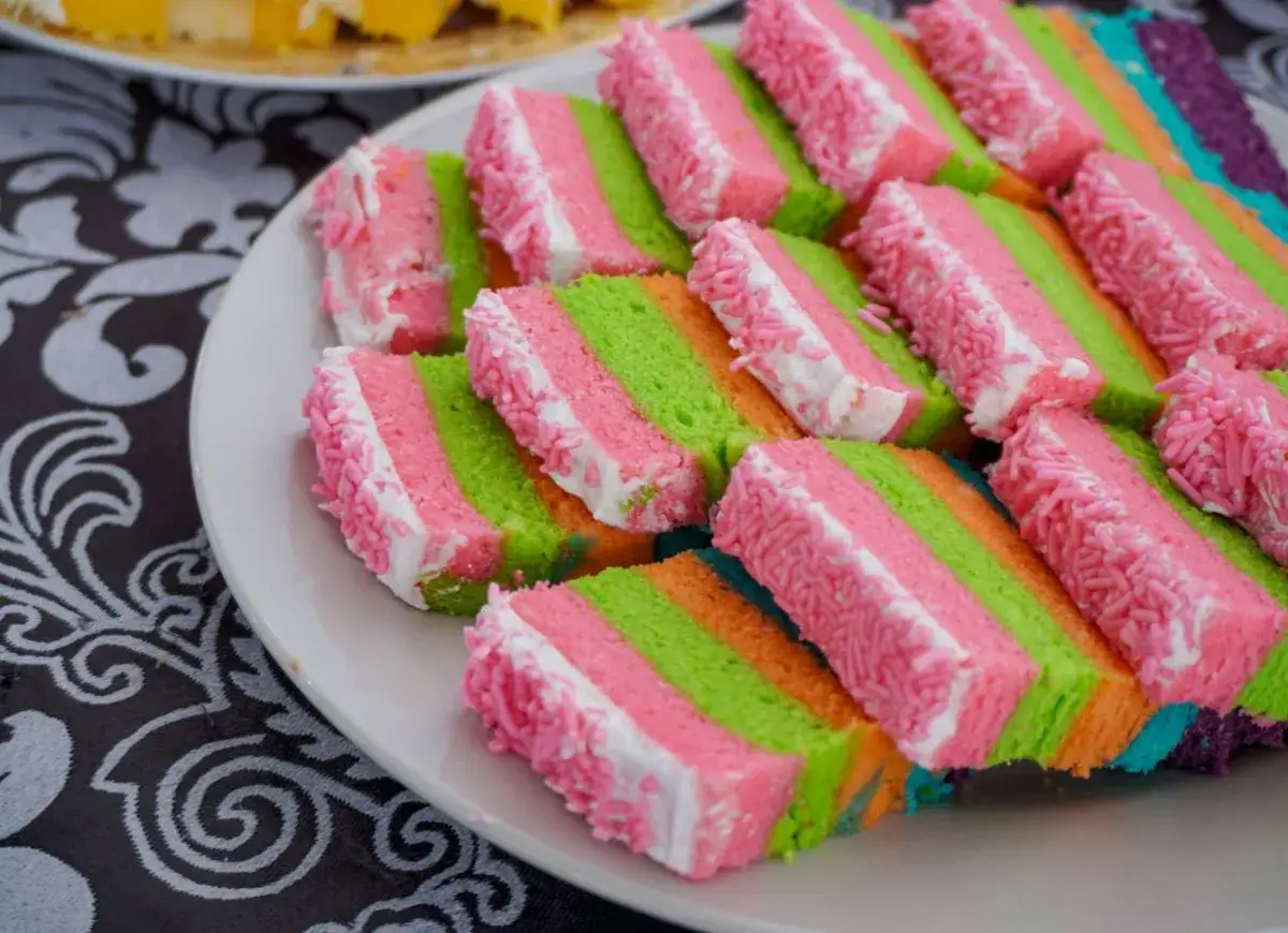 Kolorowe ciasto dla dzieci do przedszkola, pokrojone w paski różowe, zielone i pomarańczowe, ozdobione białym kremem i różową posypką.