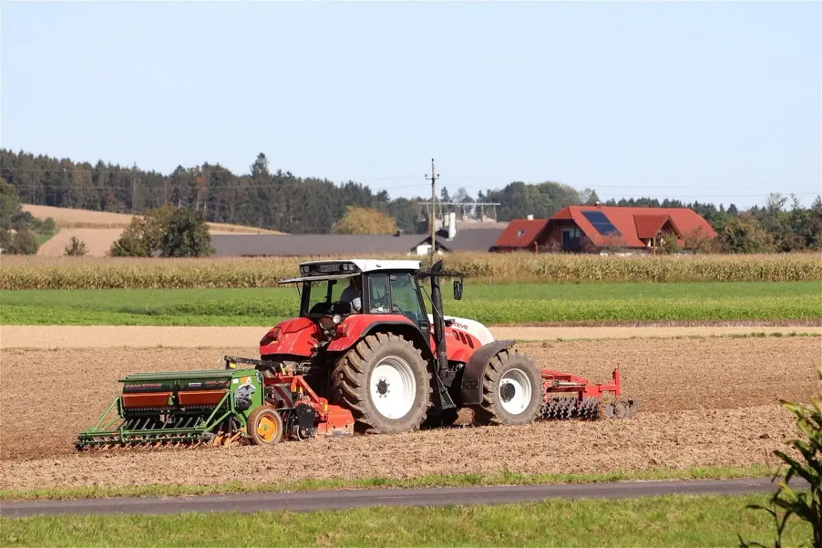 Czerwony traktor z siewnikiem na polu, w tle domy i las.