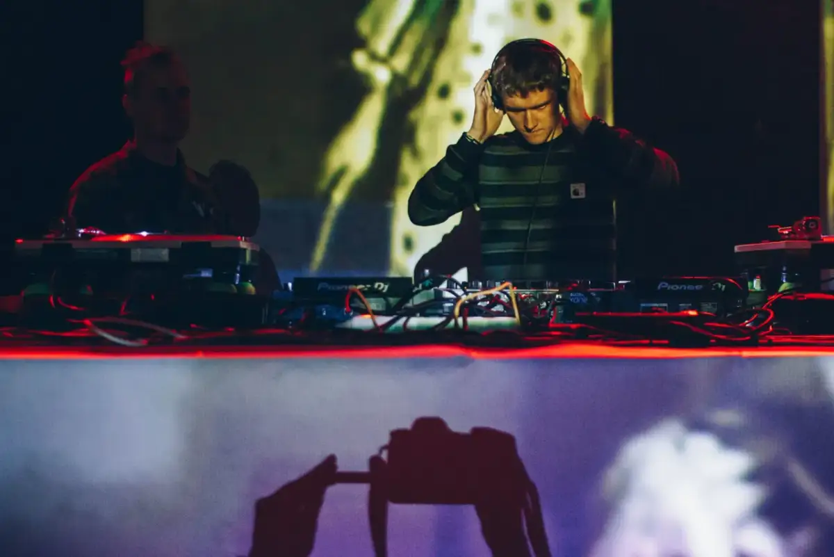 DJ w słuchawkach przy konsoli Pioneer DJ, w tle inny DJ i projekcja.