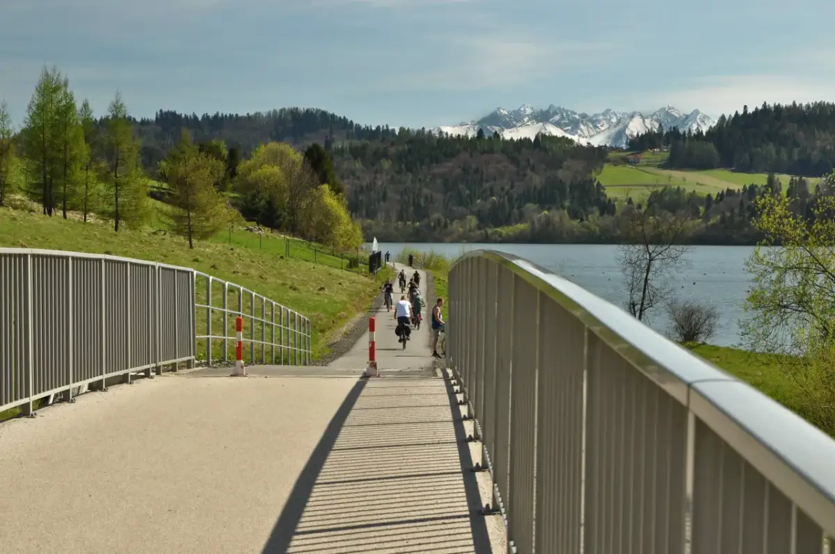 Rowerzyści na malowniczej trasie rowerowej w Czorsztynie, z widokiem na jezioro i ośnieżone szczyty Tatr.