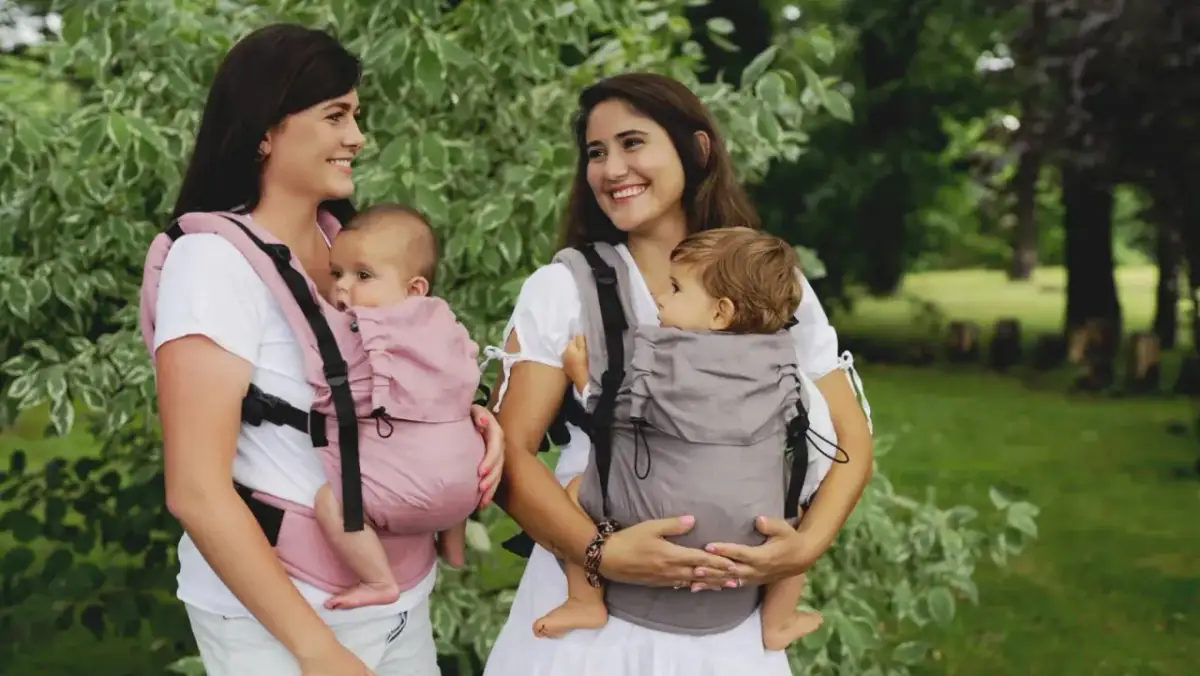 Dwie mamy z niemowlętami w nosidełkach. Fizjoterapeuta poleciłby takie nosidełko dla prawidłowego rozwoju dziecka.