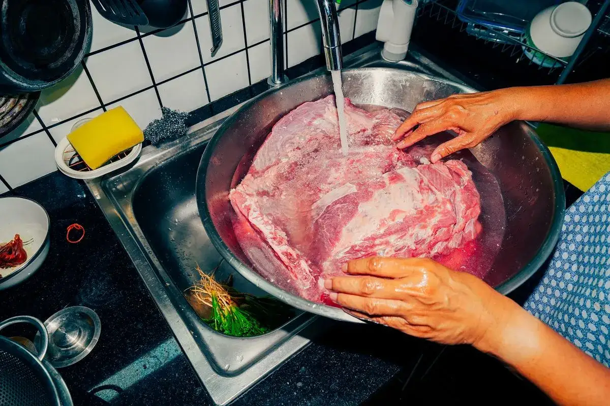 Ręce płuczą duży kawałek wieprzowiny pod bieżącą wodą. To ważny krok, by dowiedzieć się, ile gotować wieprzowinę.