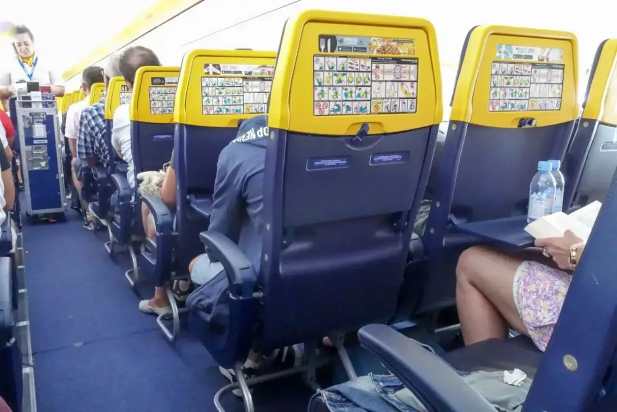 Wnętrze samolotu Ryanair. Widok na rząd siedzeń z żółtymi zagłówkami i instrukcjami bezpieczeństwa. Numeracja miejsc w samolocie Ryanair jest widoczna.