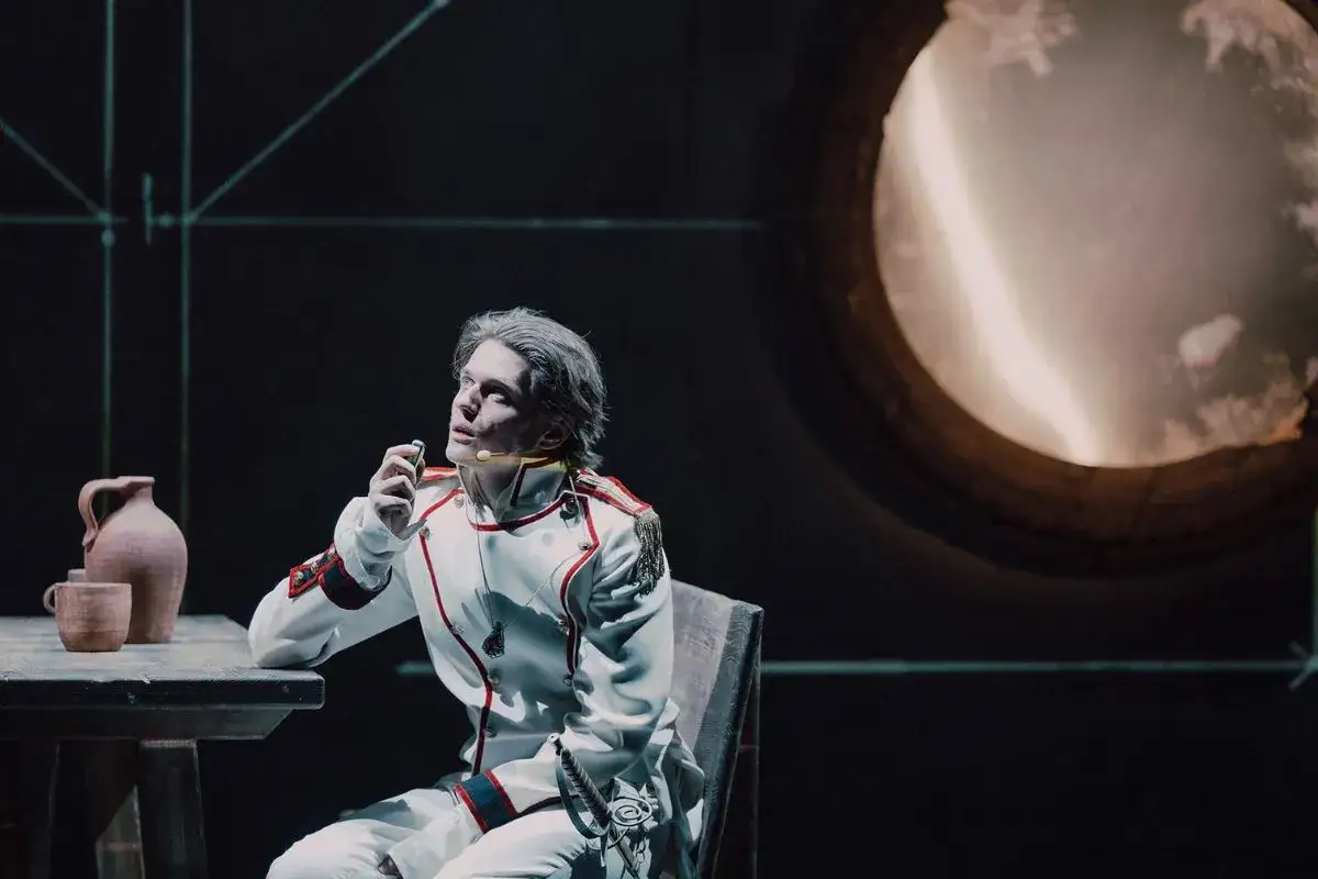 Aktor w białym mundurze siedzi przy stole, trzymając w dłoni mały przedmiot.