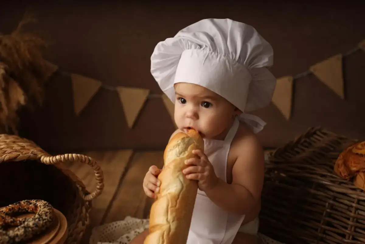 Niemowlę w czepku kucharskim gryzie bagietkę, siedząc obok koszyka z pieczywem.