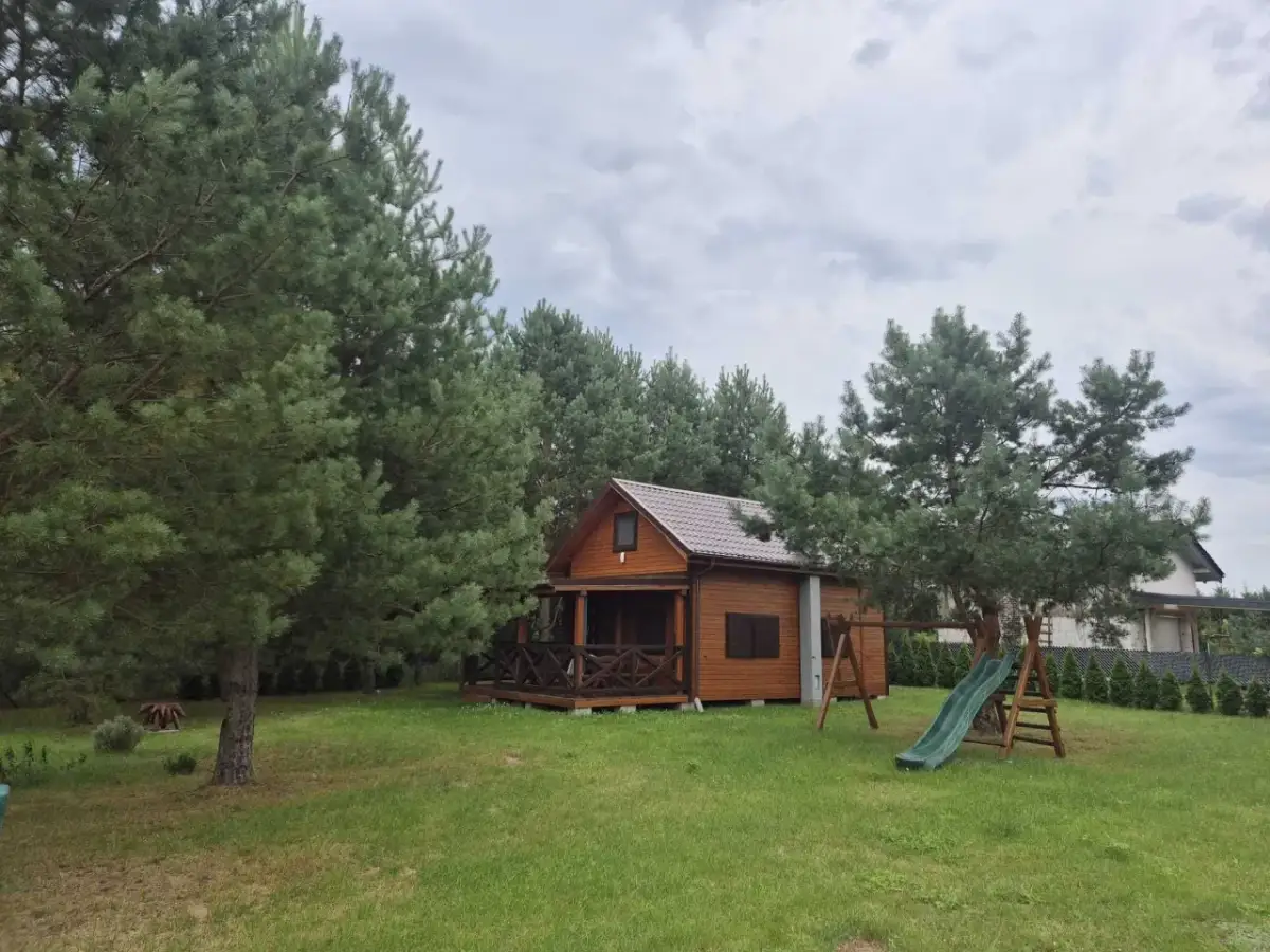 Drewniany domek letniskowy otoczony drzewami, z werandą i zjeżdżalnią.