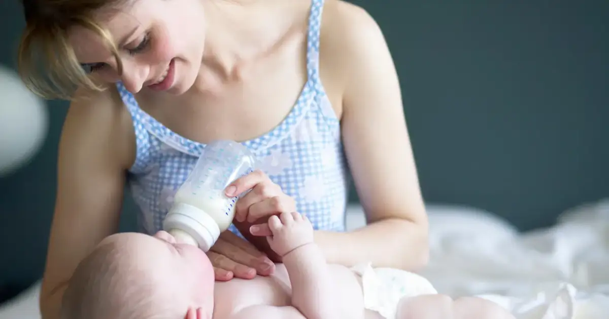 Uśmiechnięta mama karmi butelką noworodka. Warto wiedzieć, ile powinien jeść dwutygodniowy noworodek, by zapewnić mu prawidłowy rozwój.