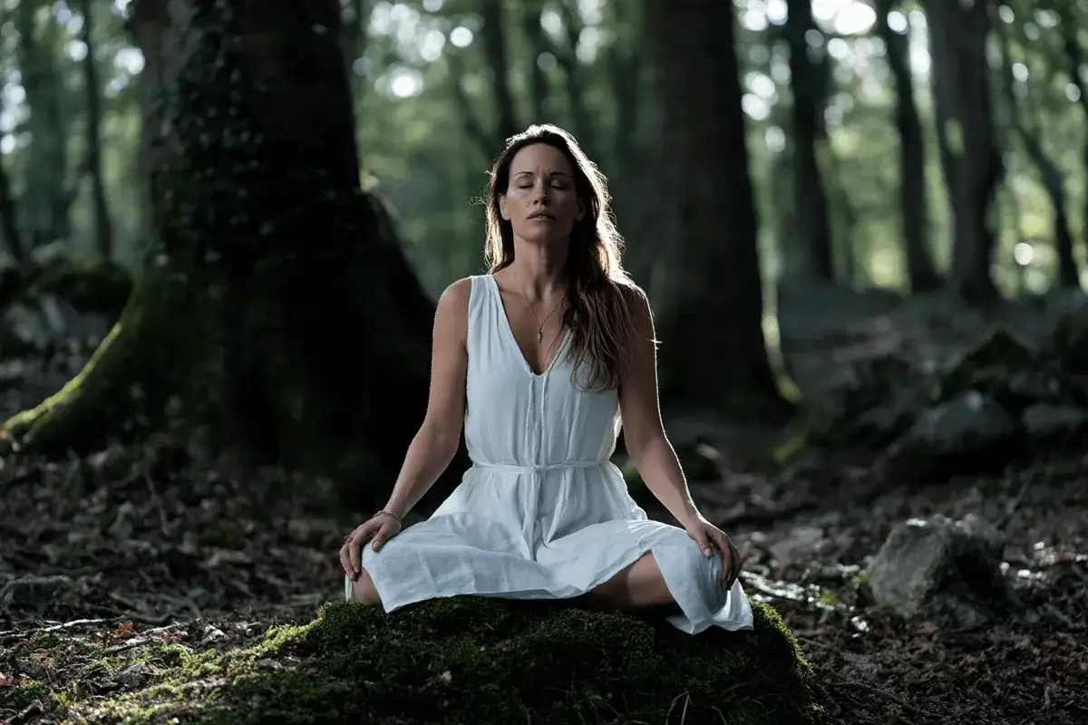 Kobieta w białej sukience medytuje w lesie na omszałym kamieniu.