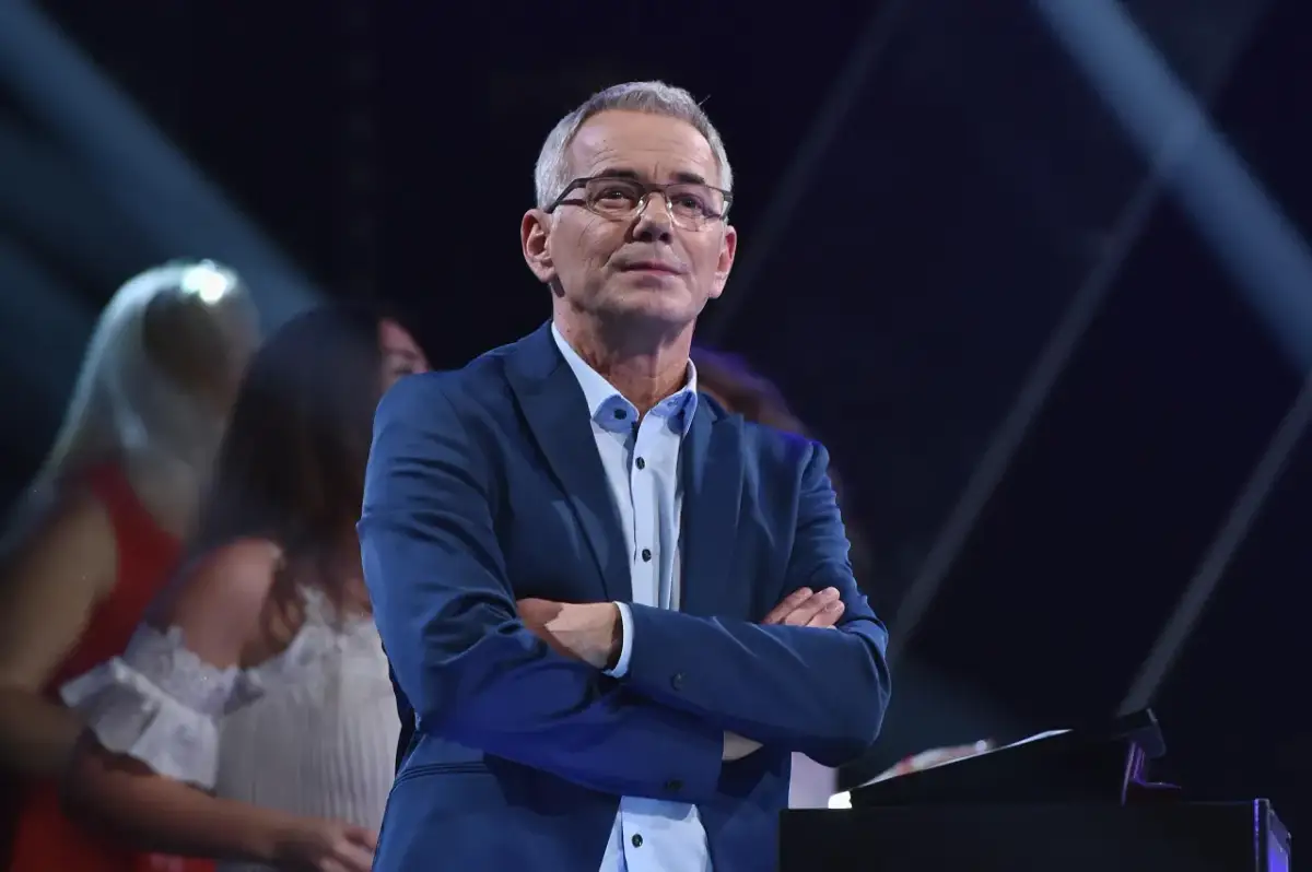 Robert Janowski w garniturze, z rękami skrzyżowanymi na piersi, na tle sceny. Czy to właśnie powód, dlaczego Robert Janowski naprawdę odszedł z Jaka to melodia po latach?