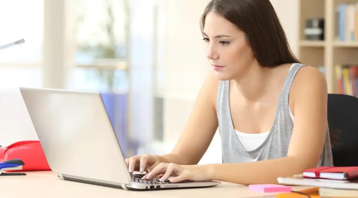 Dziewczyna pracuje na laptopie, zastanawiając się, co to jest wpisowe. Na biurku leżą zeszyty i czerwony piórnik.