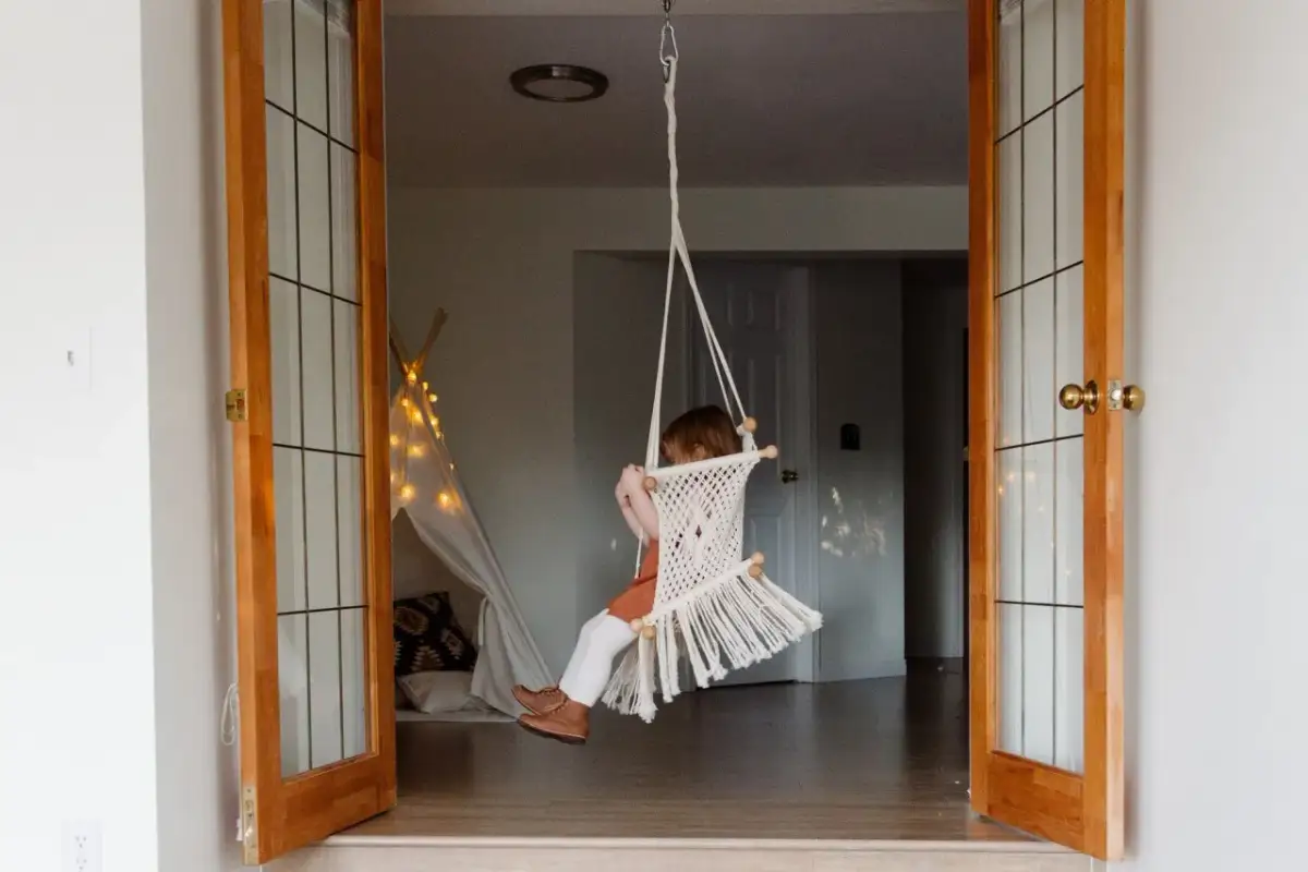 Dziecko buja się na huśtawce z makramy, wiszącej w futrynie. Pomysł na to, jak zamontować huśtawkę w futrynie.