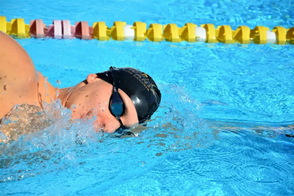 Pływak w czepku i okularach pokonuje wodę, pokazując, jak dobrze pływać kraulem. W tle widać żółte i różowe boje.