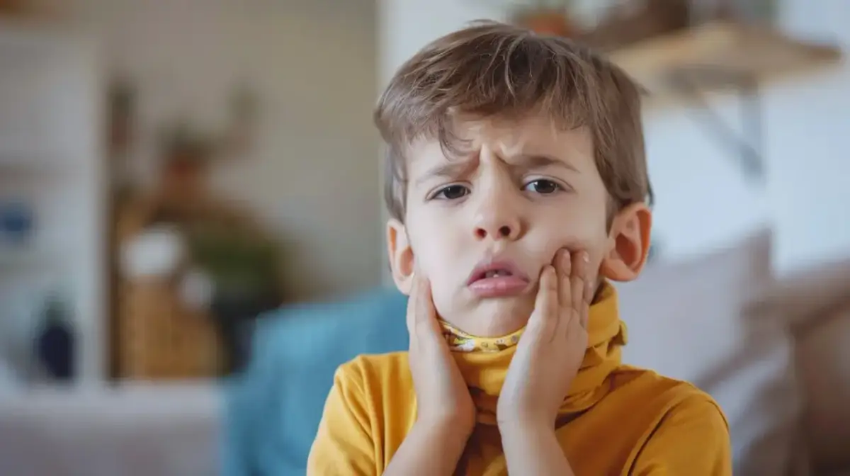 Chłopiec z grymasem na twarzy, trzymający się za policzki. Może to być objaw bólu gardła spowodowanego przez paciorkowca.
