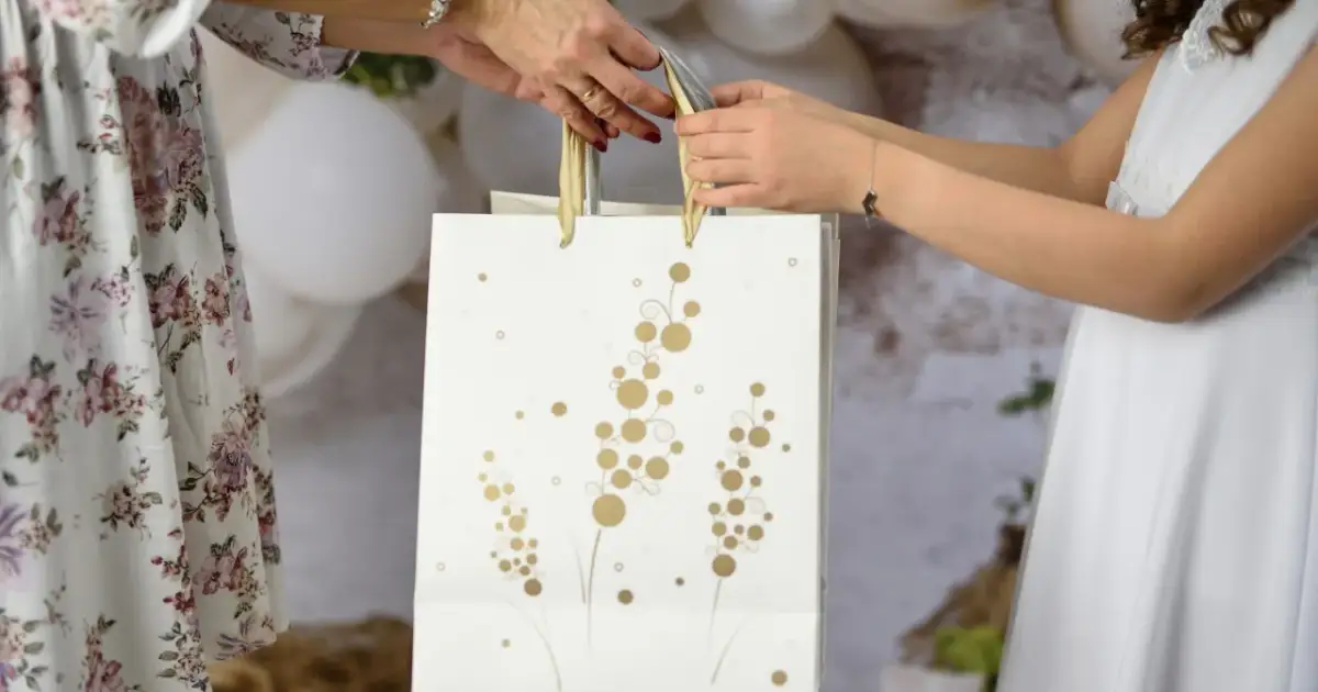 Dwie kobiety przekazują sobie białą torbę prezentową z ozdobnymi złotymi wzorami.