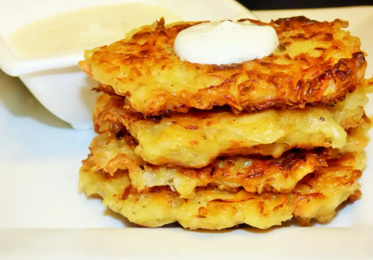 Złociste placki z kapusty kiszonej, ułożone w stosik, zwieńczone kleksem śmietany. Obok sos.