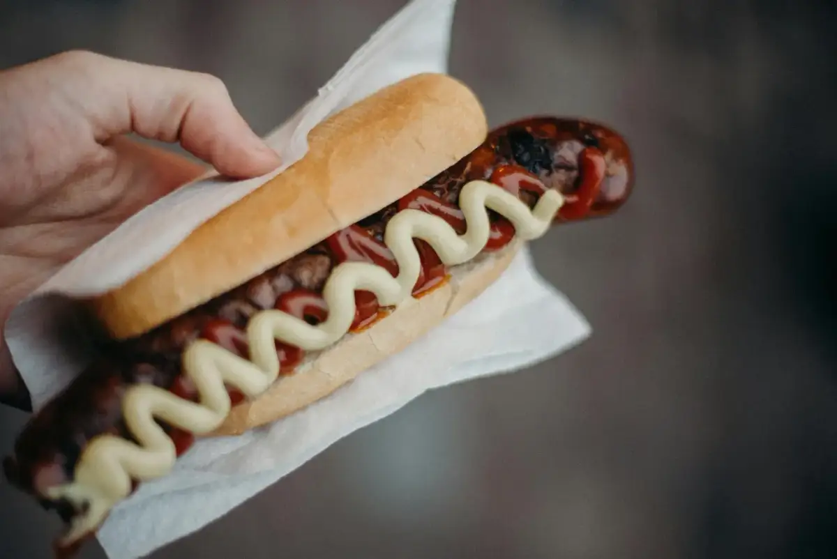 Kabanos w bułce z keczupem i musztardą, trzymany w dłoni.
