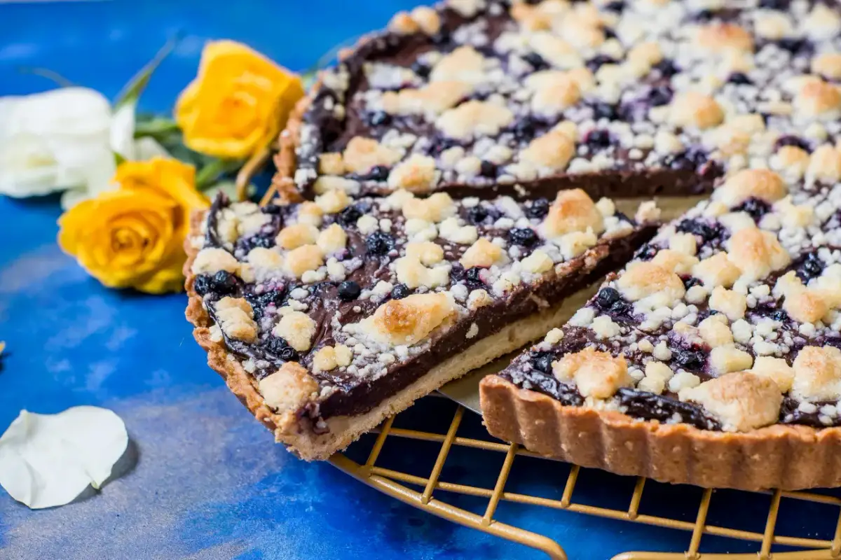 Prosta tarta z budyniem czekoladowym i jagodami, posypana kruszonką. Obok żółte róże.