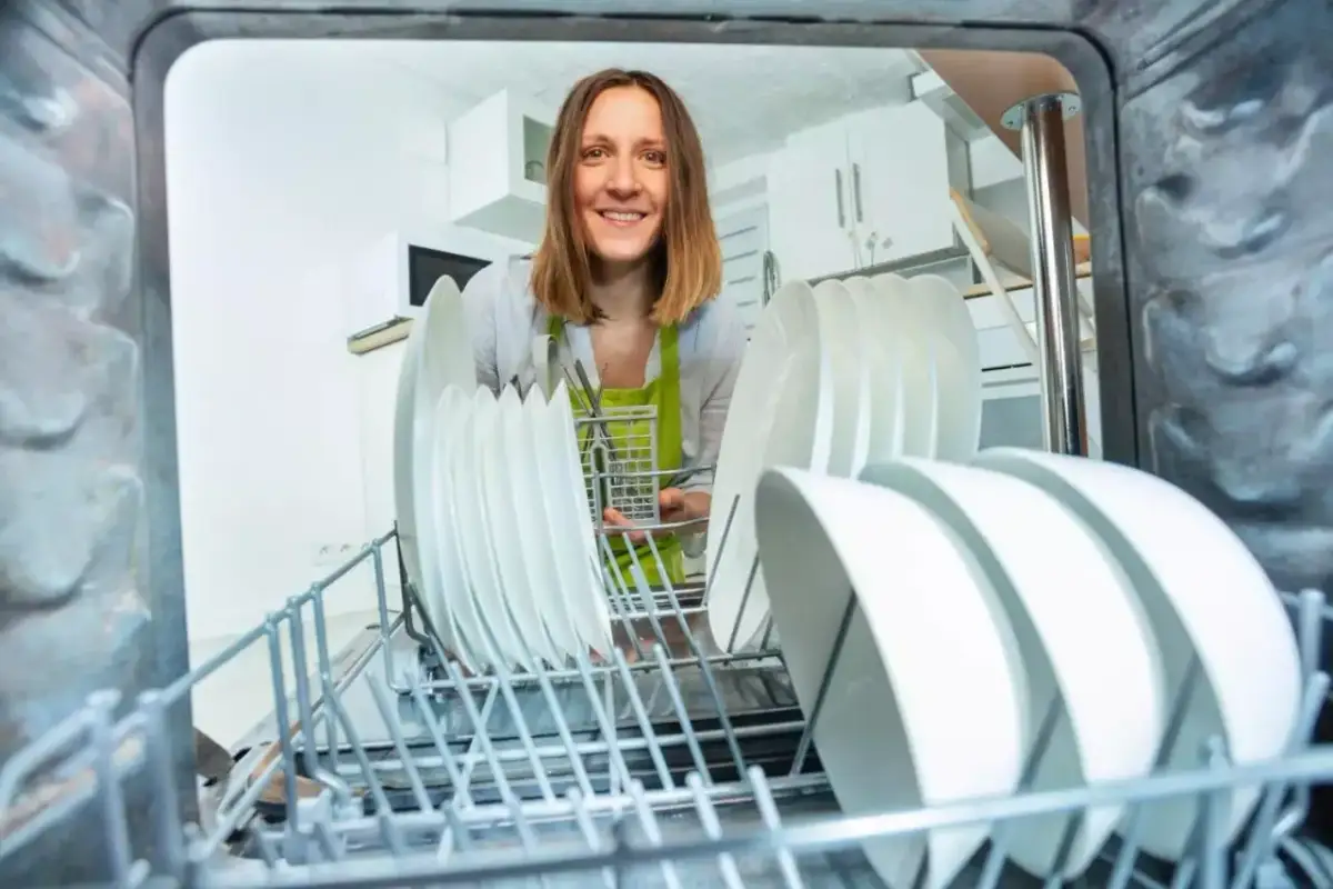 Uśmiechnięta kobieta wkłada naczynia do zmywarki. Szukasz, czym zastąpić sól do zmywarki?