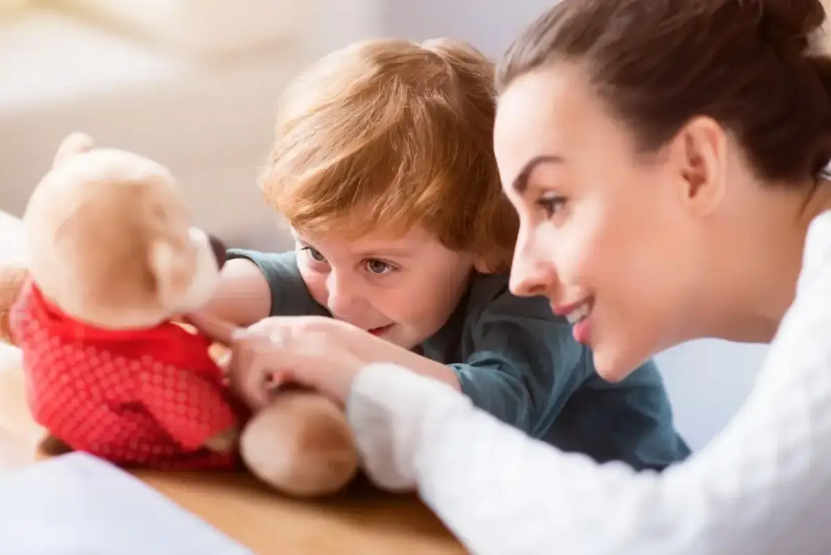 Uśmiechnięta opiekunka bawi się z dzieckiem pluszowym misiem. Zastanawiasz się, ile bierze opiekunka do dzieci za taką troskę?