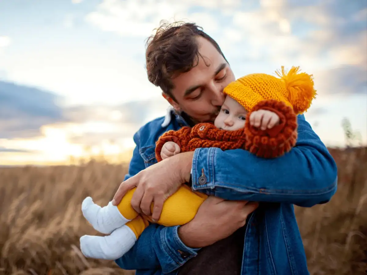 Ojciec obejmuje i całuje dziecko ubrane w żółtą czapkę i pomarańczowy sweterek na tle pola.