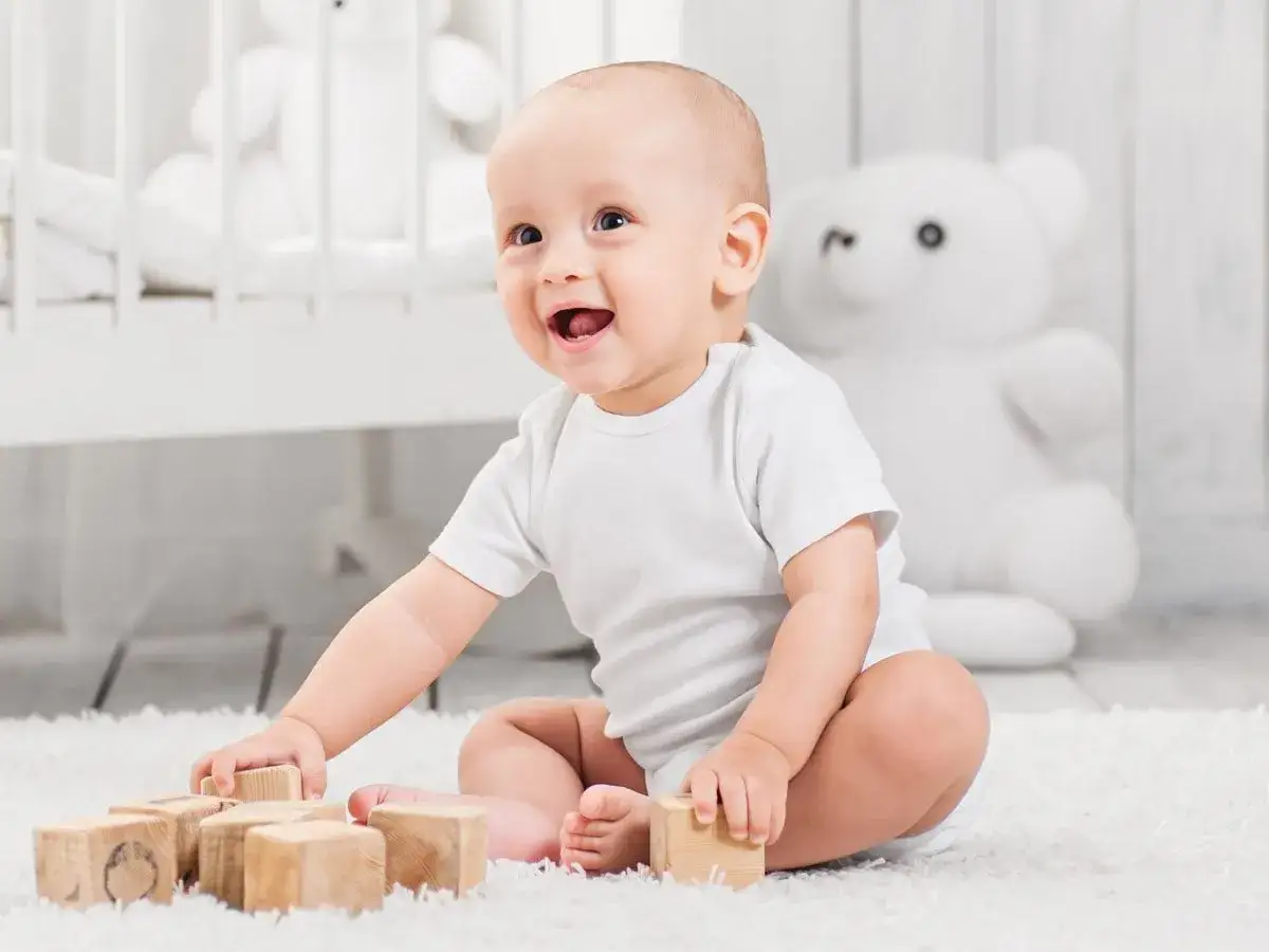 Uśmiechnięte niemowlę w białym body bawi się drewnianymi klockami na dywanie.
