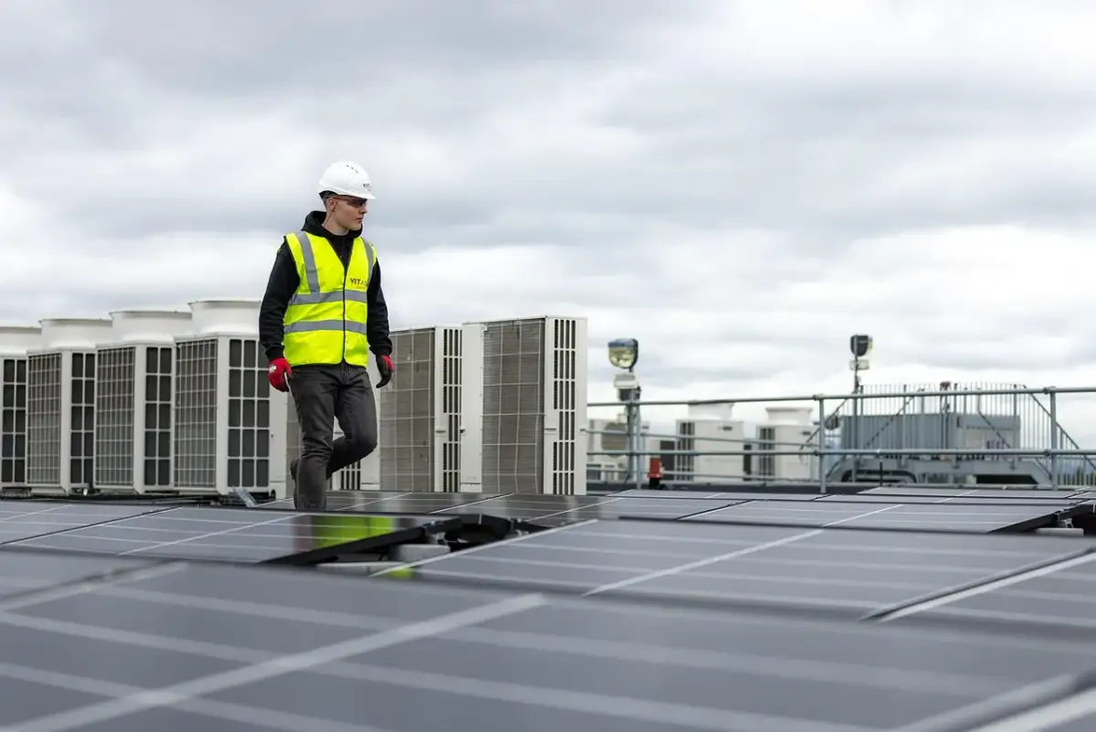 Pracownik w kamizelce odblaskowej przechodzi obok rzędu klimatyzatorów na dachu, gdzie instalowane są panele słoneczne. sofar solar polska.