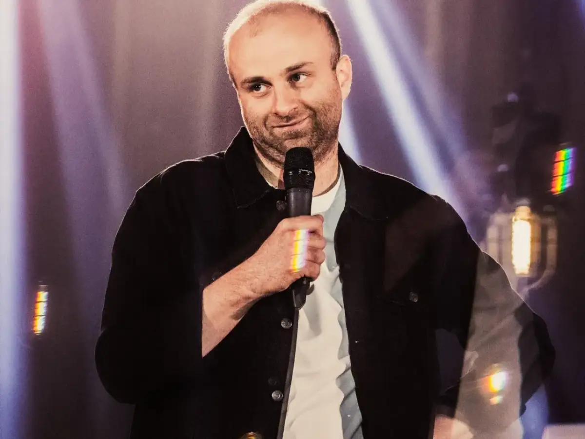 Marcin Zbigniew Wojciech w swoim stand-upie, trzyma mikrofon, a światła sceniczne tworzą tęczowe refleksy.