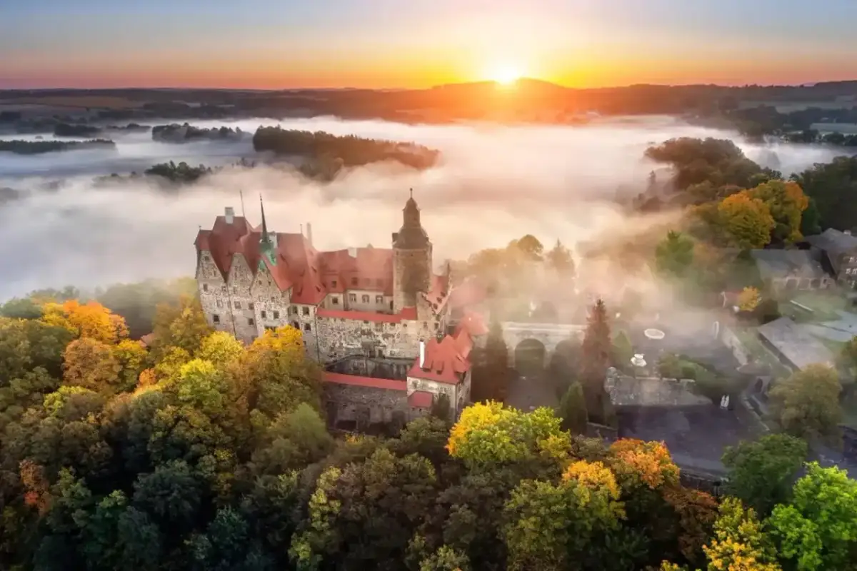 Zamek Książ otoczony jesiennymi drzewami, wyłaniający się z porannej mgły. Słońce wschodzi nad wzgórzami, malując niebo na pomarańczowo.
