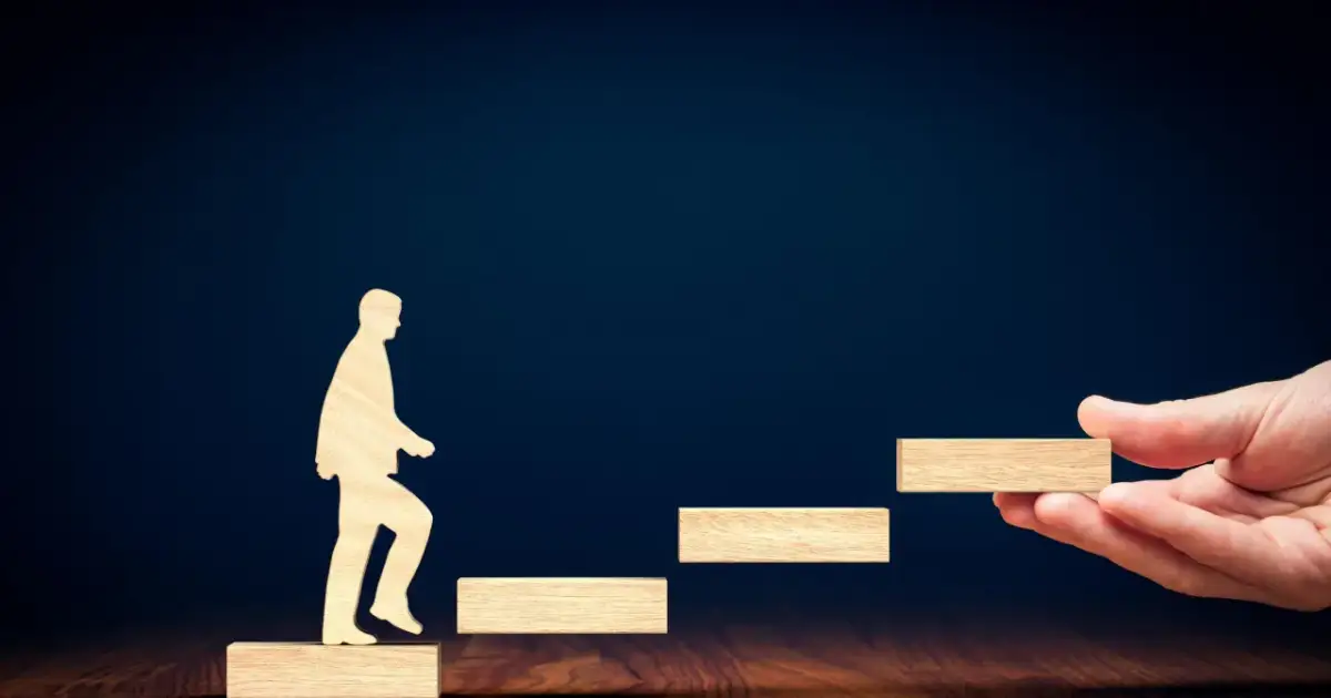 Drewniana postać wchodzi po schodkach, a dłoń podaje kolejny stopień.