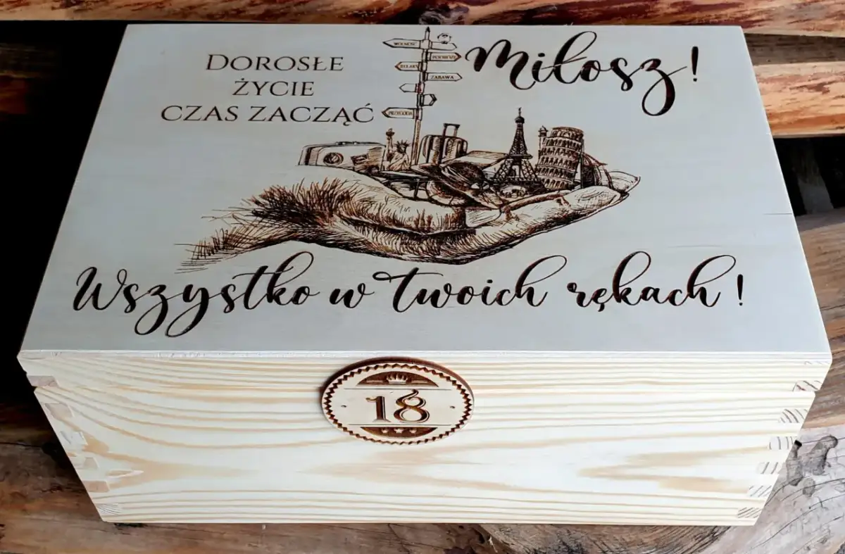 Drewniana skrzynka z grawerem na 18 urodziny od chrzestnej. Na wieczku napis "Dorosłe życie czas zacząć, Miłosz! Wszystko w Twoich rękach!" i grafika dłoni z symbolami podróży.