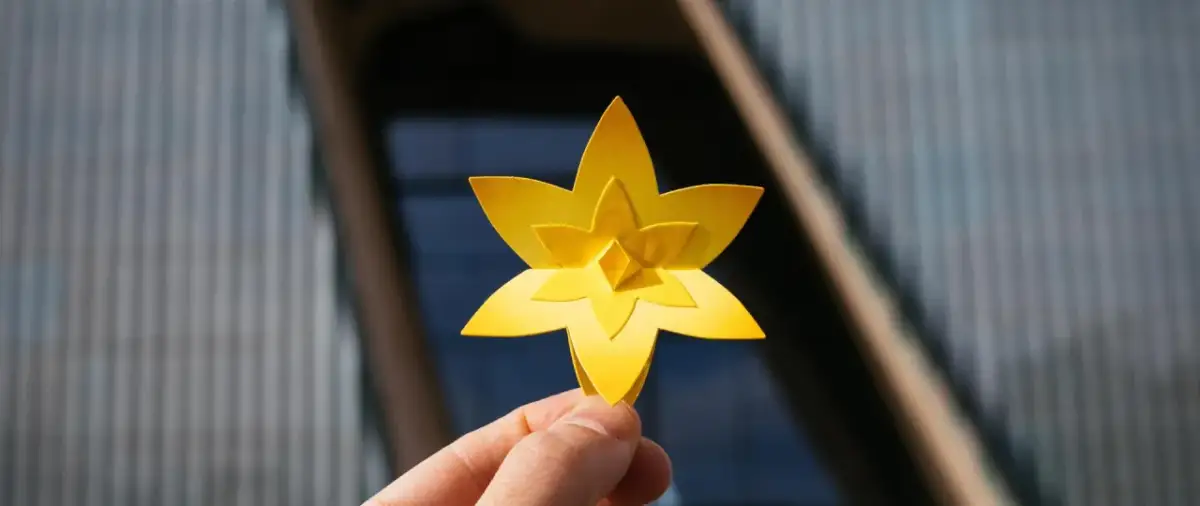 Żółty, papierowy żonkil, symbolizujący nadzieję i nowy początek, trzymany na tle nowoczesnego budynku.