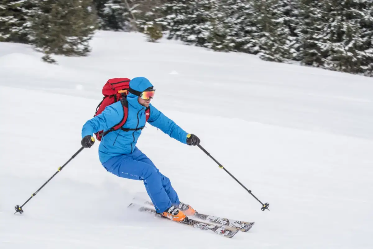 Narciarz w niebieskiej kurtce i goglach zjeżdża po ośnieżonym stoku, niosąc czerwony plecak. To prawdziwy miłośnik ski tury!