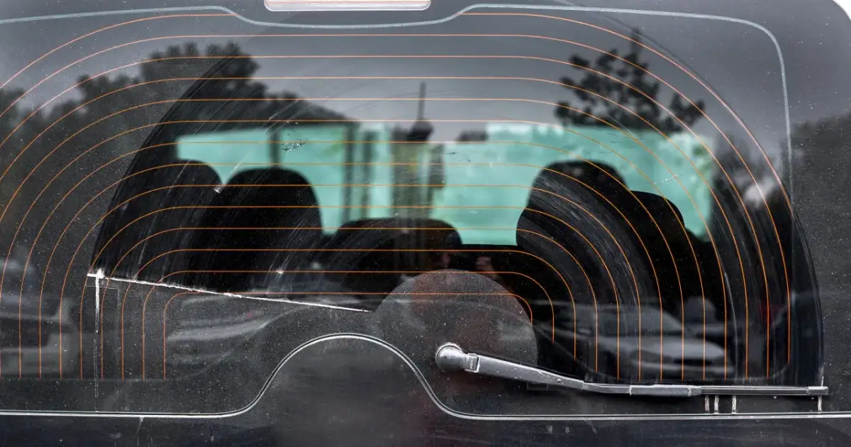 Widok tylnej szyby samochodu z widocznymi liniami ogrzewania. Widać wycieraczkę i odbicia drzew.