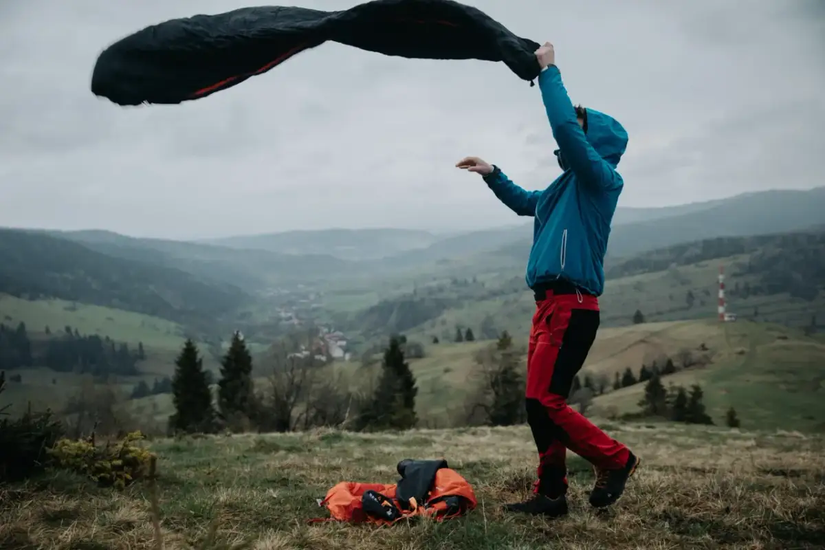 Osoba w niebieskiej kurtce i czerwonych spodniach rozkłada śpiwór puchowy czy syntetyczny na tle górskiego krajobrazu.